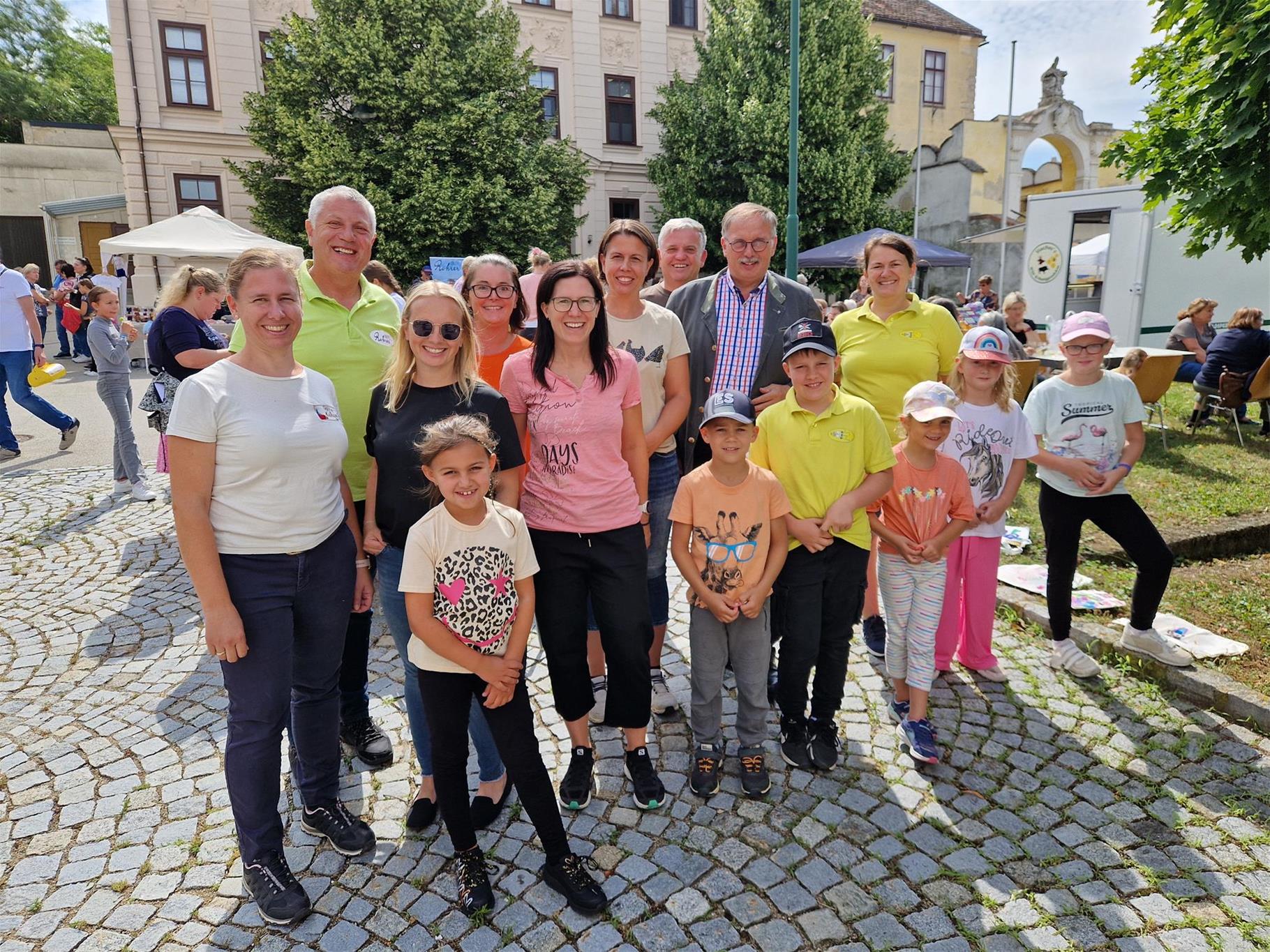 Auf dem Bild ist Bürgermeister Hogl mit Organisatoren des Schmankerlmarkts und jungen Besuchern mit lachenden Gesichtern zu sehen