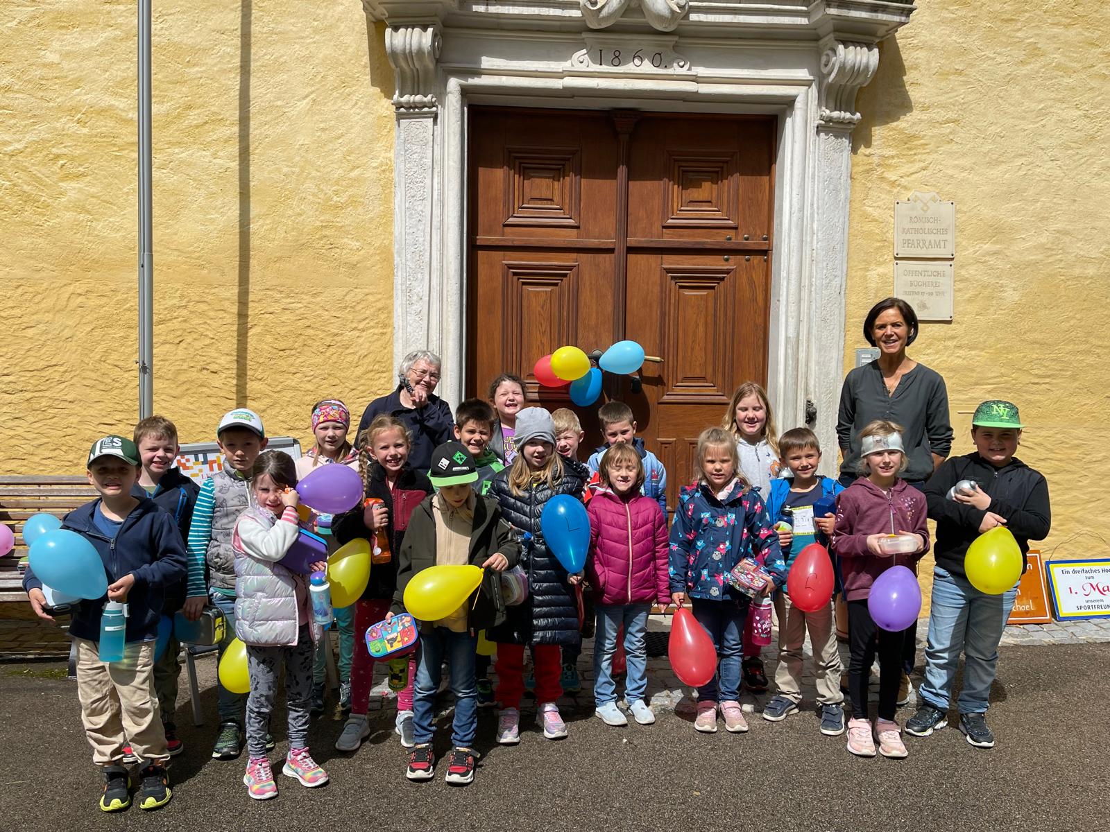 Auf dem Bild lachen Lisl Tradinik, Marianne Roher und die Erstklässler mit Luftballons in der Hand
